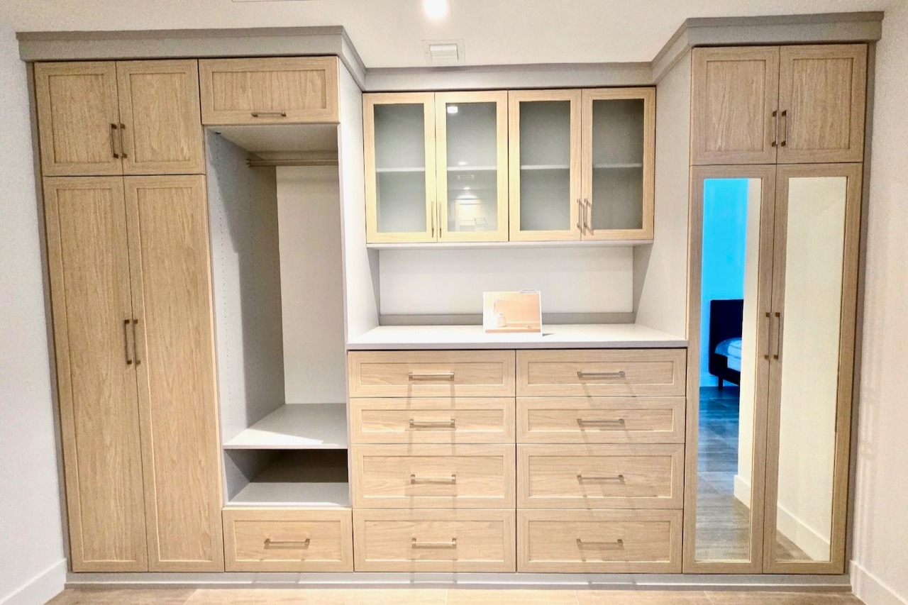 Walk-In Closet in Sunset Mist with Glass Door Inserts Walk-in closet in Sunset Mist finish with natural doors and drawer faces, featuring satin and mirrored glass door inserts for a stylish and functional storage solution.
