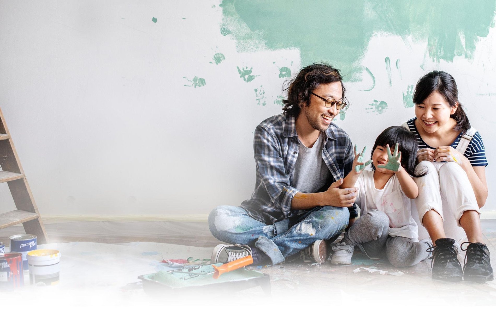 Photo of a smiling family sitting on the floor of a new home, their small child has put handprints on the wall with wet paint