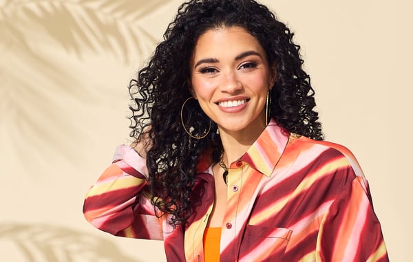 Woman is posing in front of a tan background while wearing a yellow, red and orange button down shirt.