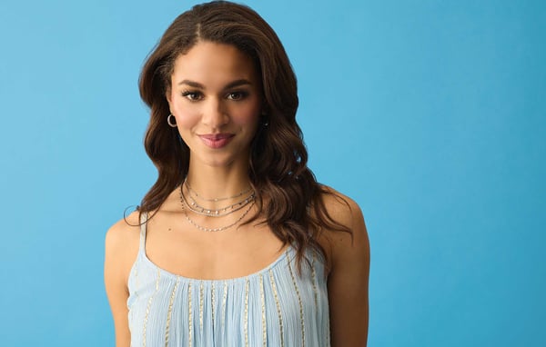 Woman wearing a light blue dress with layered gold necklaces is smiling softly in front of a blue background.