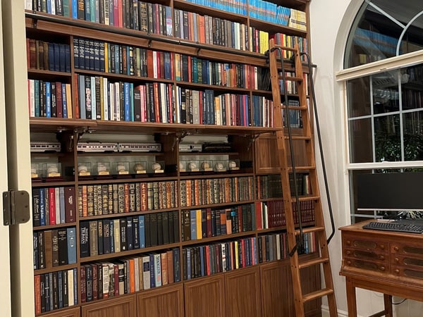 A timeless custom home library designed to showcase an extensive book collection with rich wood cabinetry, floor-to-ceiling shelving, and a rolling library ladder that brings classic character to the space. Custom home library with floor-to-ceiling wood bookshelves, rolling library ladder, lower cabinetry, and extensive book storage.