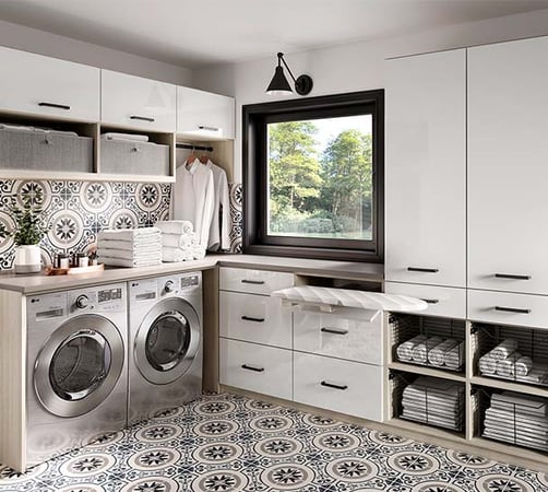 This stylish and highly functional laundry room features sleek cabinetry, bold patterned tile, and smart storage solutions. Integrated appliances, a pull-out ironing board, and open shelving keep everything organized and within reach—perfect for streamlining your daily routine. Modern laundry room with front-loading washer and dryer, white cabinetry, patterned tile flooring and backsplash, open shelving with towels and storage bins, a pull-out ironing board, and a window with a view of green trees.