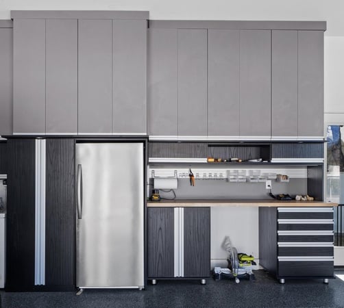 This modern garage setup combines sleek design with high-performance function. Featuring custom cabinetry in matte gray and textured black finishes, a stainless steel fridge, a built-in workbench, and tool organization solutions—this space is built for efficiency and style. Designed and installed by California Closets of the Texas Hill Country. Modern garage storage system by California Closets of the Texas Hill Country, featuring tall gray upper cabinets, black lower cabinets with metallic accents, a stainless steel refrigerator, wood workbench, tool wall organizer, and drawer units.