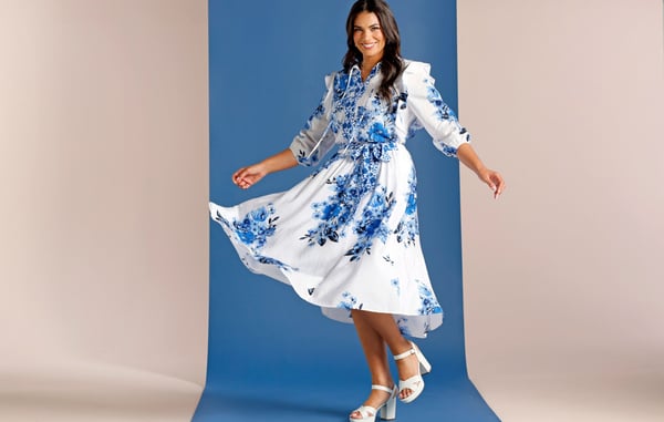 Woman wearing a plus size floral blue and white blouse with a matching plus size blue and white midi skirt and white platform heels is posing in front of a blue and tan background.