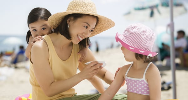Mom applying sunscreen to daugther while sitting on the beach with other daughter peeking over her shoulder