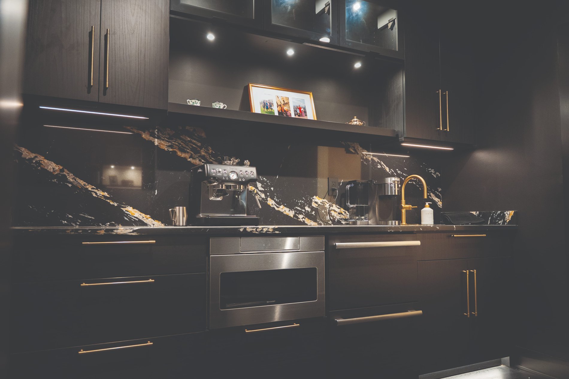 Custom Coffee Bar Cabinetry with Integrated Lighting Custom coffee bar cabinetry featuring Shadow Black cabinets, under-cabinet lighting, a coffee station, stone backsplash, and built-in storage.