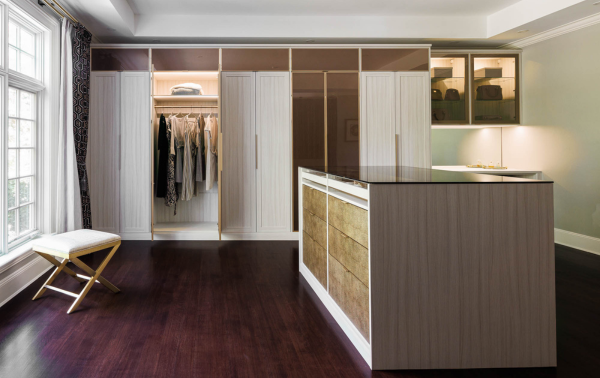 Two Toned White & Bronze Walk-in Closet California Closets Westchester NY Full Room Dressing Room Walk-in Closet with Glass Bronze Doors Closed Cabinetry and Center Island with Brown Leather Drawer Faces and Clear Bronze Glass Countertop