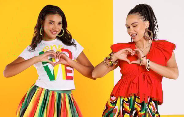 Two women wearing trendy women's clothing are posing with their hands in heart shapes.