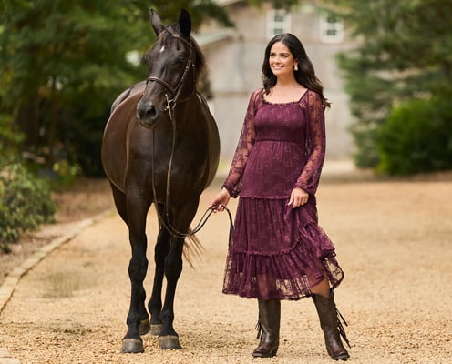 Woman is wearing a purple lace maxi dress and is standing next to a brown horse.