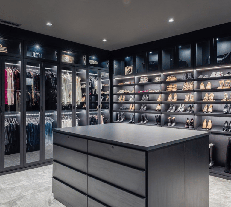 Lux Modern Primary Walk-in Closet Black walk-in closet with glass door inserts, lighting, center island and slanted shoe shelves.