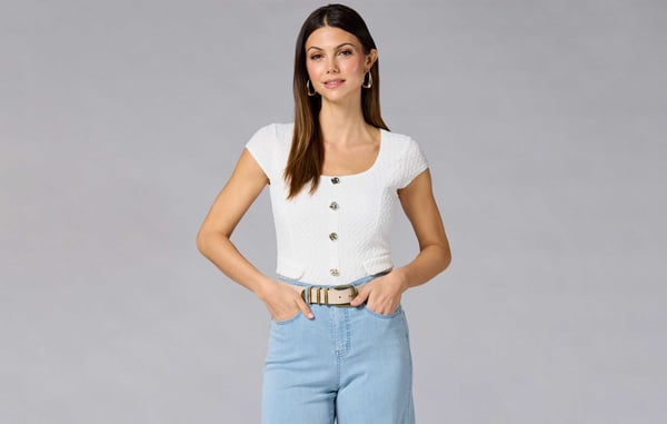 Woman wearing a white cap sleeve top with gold buttons and blue jeans is posing with her hands in her pockets in front of a gray background.