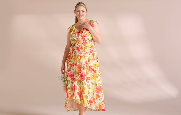 Woman is wearing a yellow, pink and orange floral flutter sleeve plus size dress, and is posing in front of a tan background.