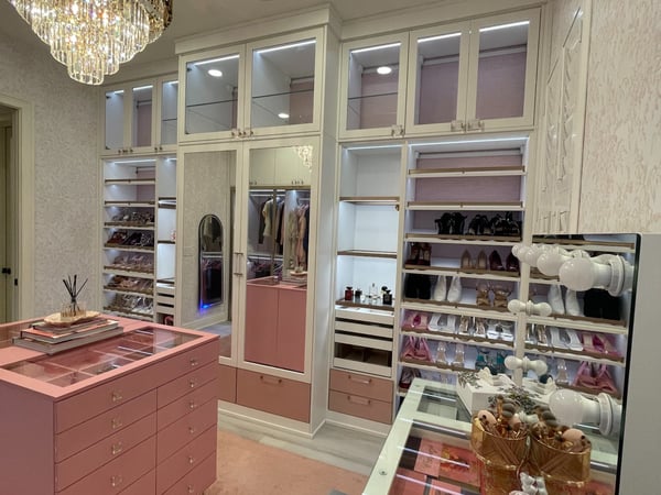 A glamorous dressing room with blush-toned cabinetry, illuminated glass displays, open wardrobe storage, and a statement island that creates a distinctly boutique-inspired experience. Luxury dressing room with blush pink cabinetry, illuminated glass cabinets, center island, shoe storage, and custom wardrobe organization.