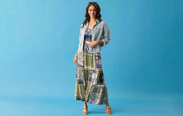 Woman is wearing a blue and green maxi dress with a denim jacket, and is posing in front of a blue background.