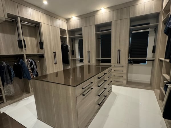 A spacious walk-in closet pairs warm wood cabinetry with a statement center island, creating generous storage for clothing, accessories, and everyday wardrobe essentials. Luxury custom walk-in closet with large center island, wood cabinetry, built-in drawers, hanging storage, and open shelving.