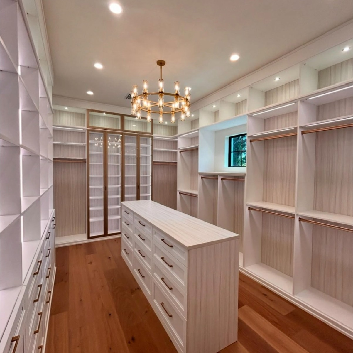 Serene Primary Walk-In Closet with Dove Finish and Modern Accents Primary walk-in closet with Dove finish, Tuscan Moon backing, aluminum frame doors, Lexington modern mitered drawers, and integrated strip LED lighting.