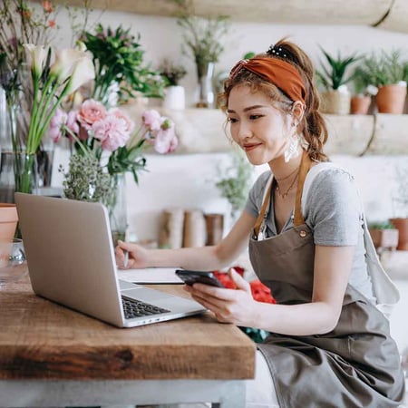 A woman in a flower shop sitting with a smartphone in her left hand and looking at a laptop computer