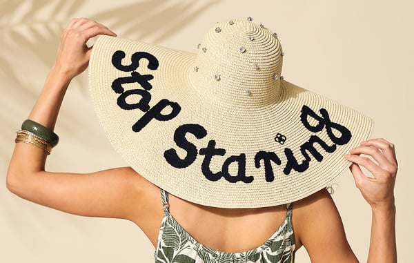 Woman is wearing a large straw sun hat that has 'Stop Staring' embroidered in black thread.