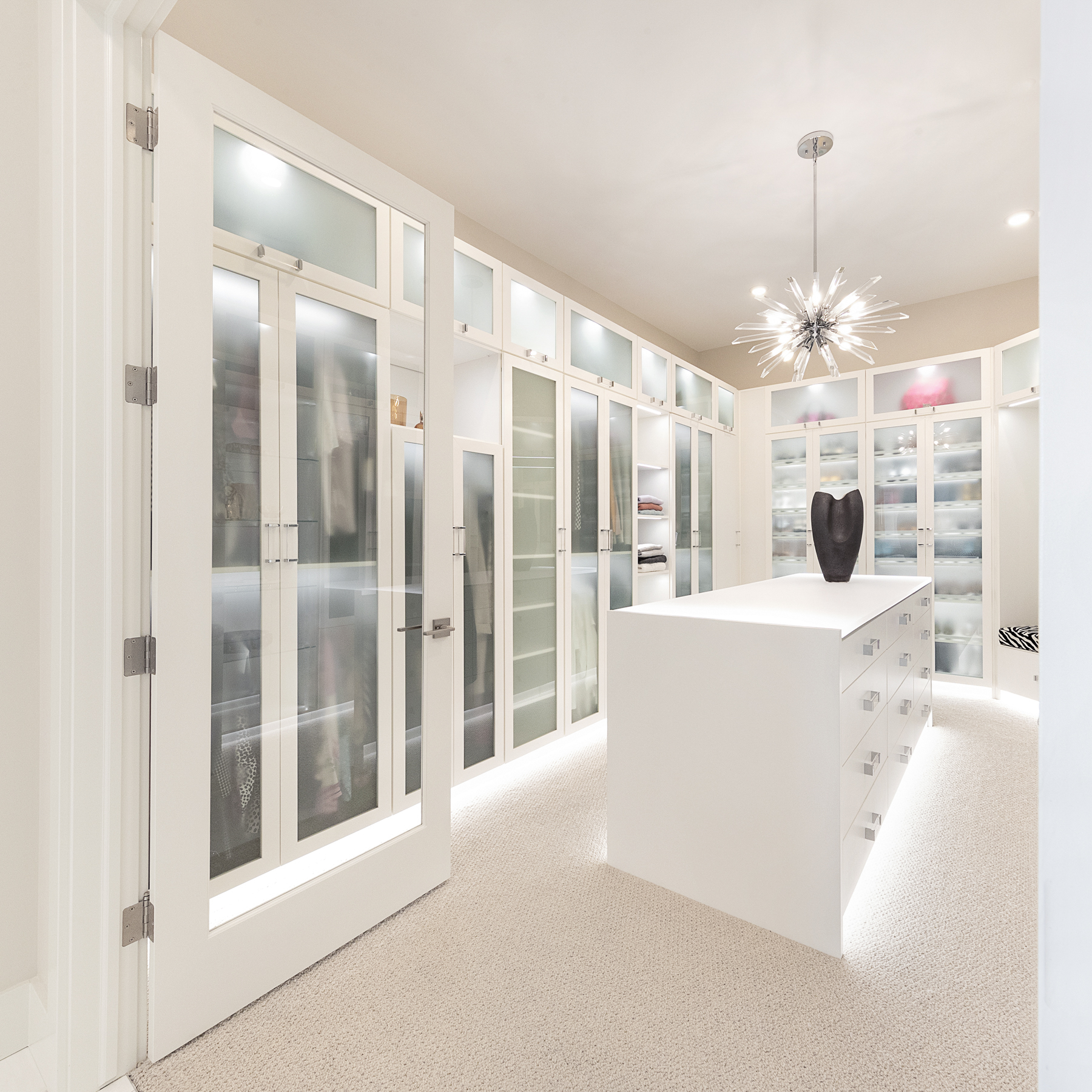 Luxe Walk-In Closet White walk-in closet with floor to ceiling glass cabinetry, center island, and under-cabinet lighting by California Closets Edmonton, Alberta.