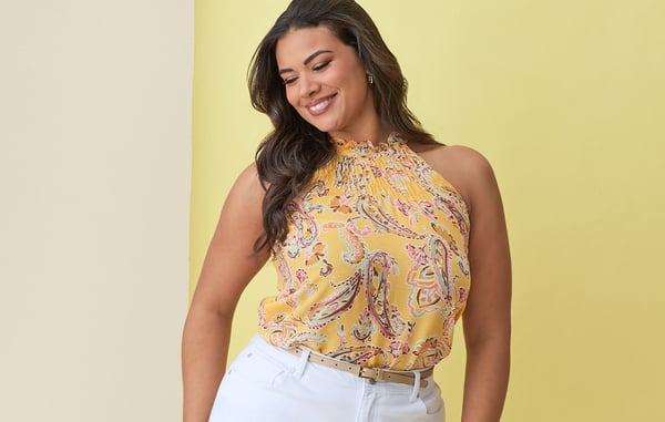 Woman wearing a plus size yellow paisley halter top and white curvy jeans is smiling and looking down in front of a yellow background.
