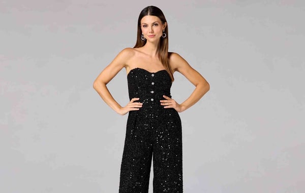Woman is posing with her hands on her hips in front of a gray background and is wearing a sleeveless black sparkly jumpsuit.