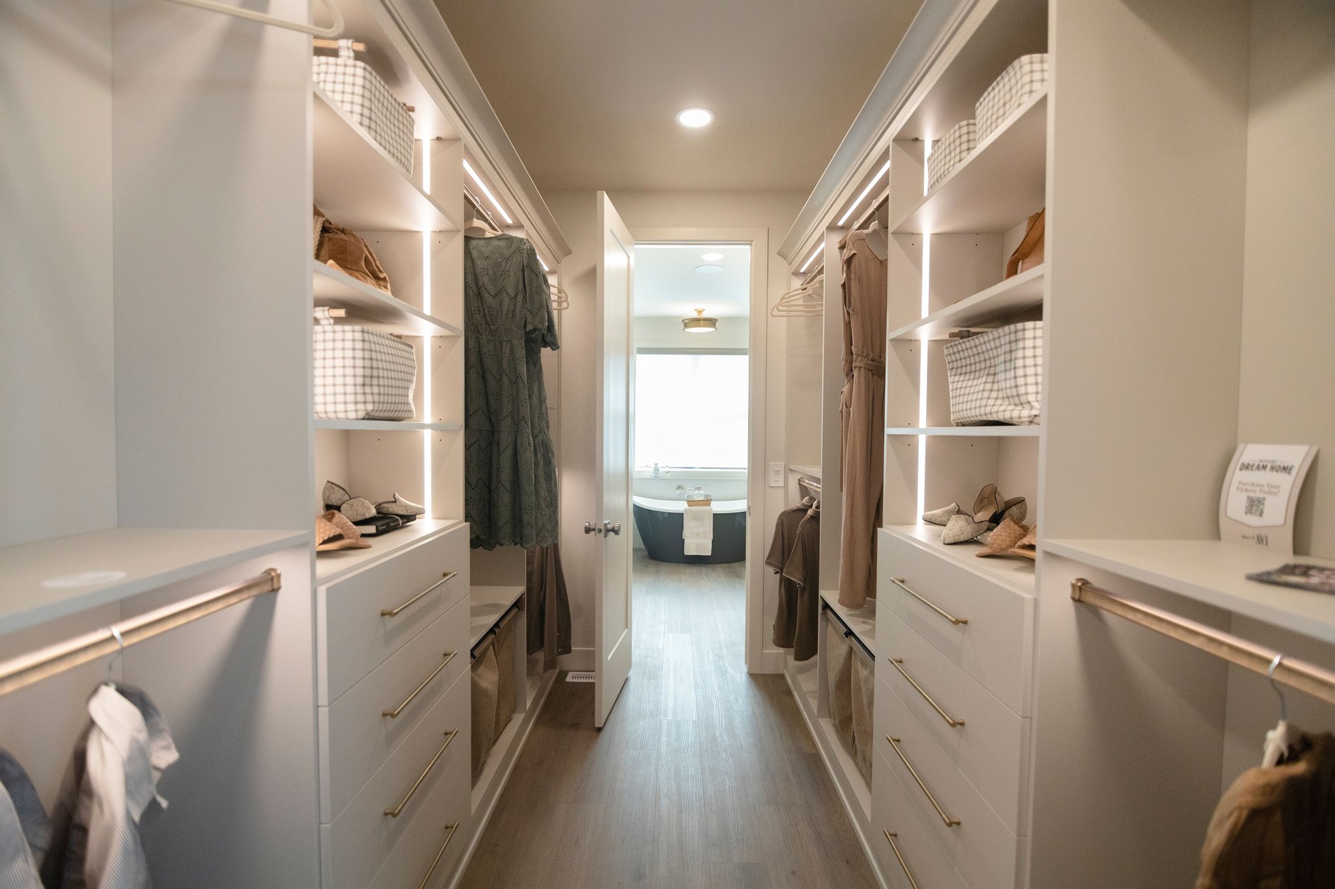 Walk Through Primary Closet Walk through closet in a warm off white finish, gold hardware, pull out hampers, and lighting.