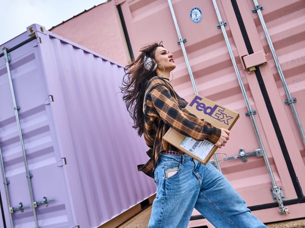 A customer carrying a FedEx flat-rate shipping package, walking past large shipping containers.