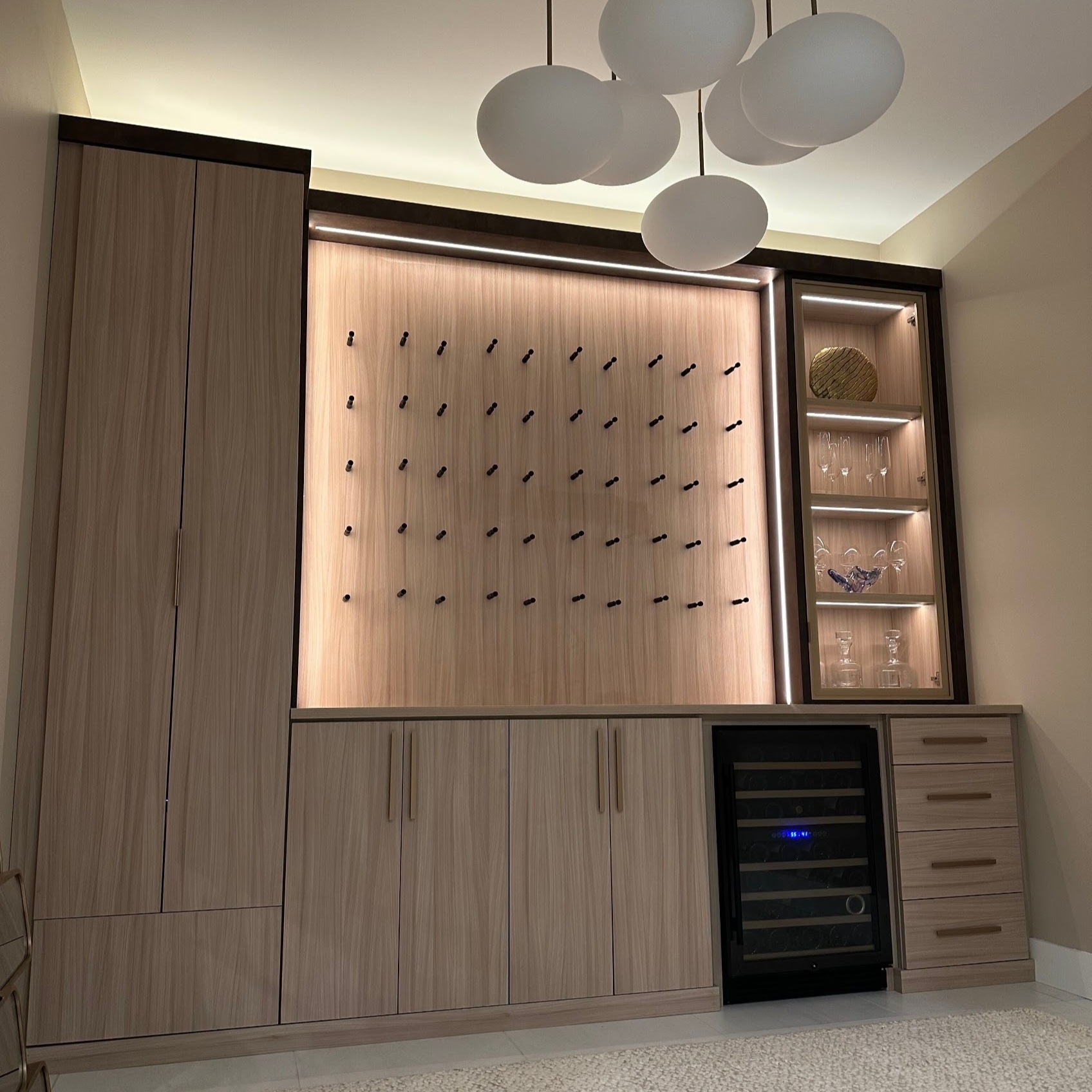 Wine Display Wall Custom wine wall with illuminated bottle display, wood cabinetry, built-in wine refrigerator, drawers, and display shelving.