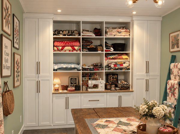 A colorful custom storage solution designed to keep creative supplies beautifully organized, combining generous shelving, drawers, countertop space, and concealed cabinetry. Custom craft room storage with built-in white cabinetry, open shelving, drawers, countertop workspace, and organized supplies.