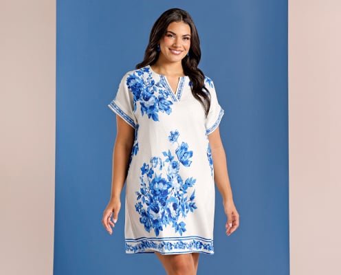 Woman is wearing a plus size blue and white floral mini shift dress and posing in front of a blue background.