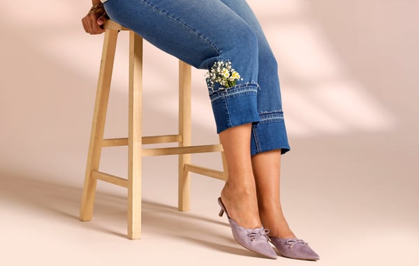 Woman is wearing plus size cropped boyfriend jeans with flowers sticking out of the hem, and posing by sitting on a wooden stool in front of a tan background.