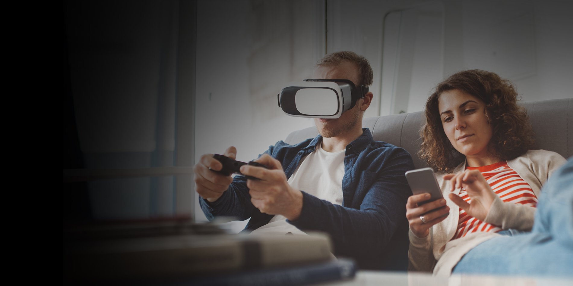 Two people on a couch using Optimum high-speed internet connected devices. One plays video games with a virtual reality headset and the other uses a smartphone.