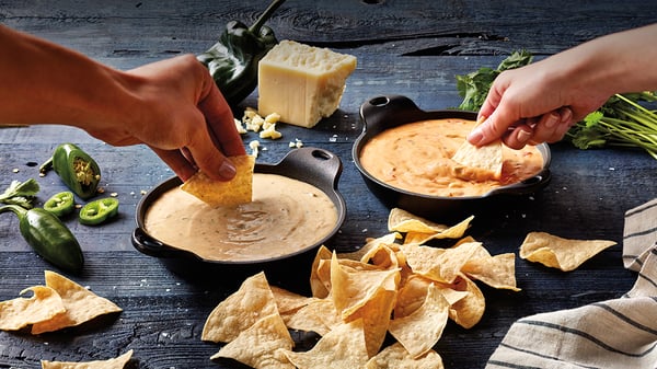 On the left hand dipping a chip into Qdoba's 3-Cheese Queso & another hand on the right dipping a chip into Quoba's Queso Diablo with a stack of chips lining the foreground.