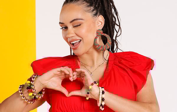 Woman wearing a red short sleeve blouse and bold statement jewelry is winking at the camera and posing with her hands in the shape of a heart.