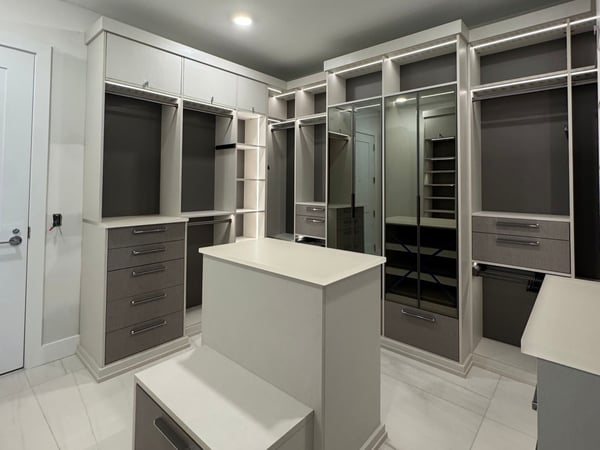 A spacious custom walk-in closet featuring a sophisticated neutral palette, generous wardrobe storage, glass-front cabinetry, drawers, and a center island for added organization. Luxury gray walk-in closet with center island, glass-front cabinetry, hanging storage, drawers, and custom shelving.