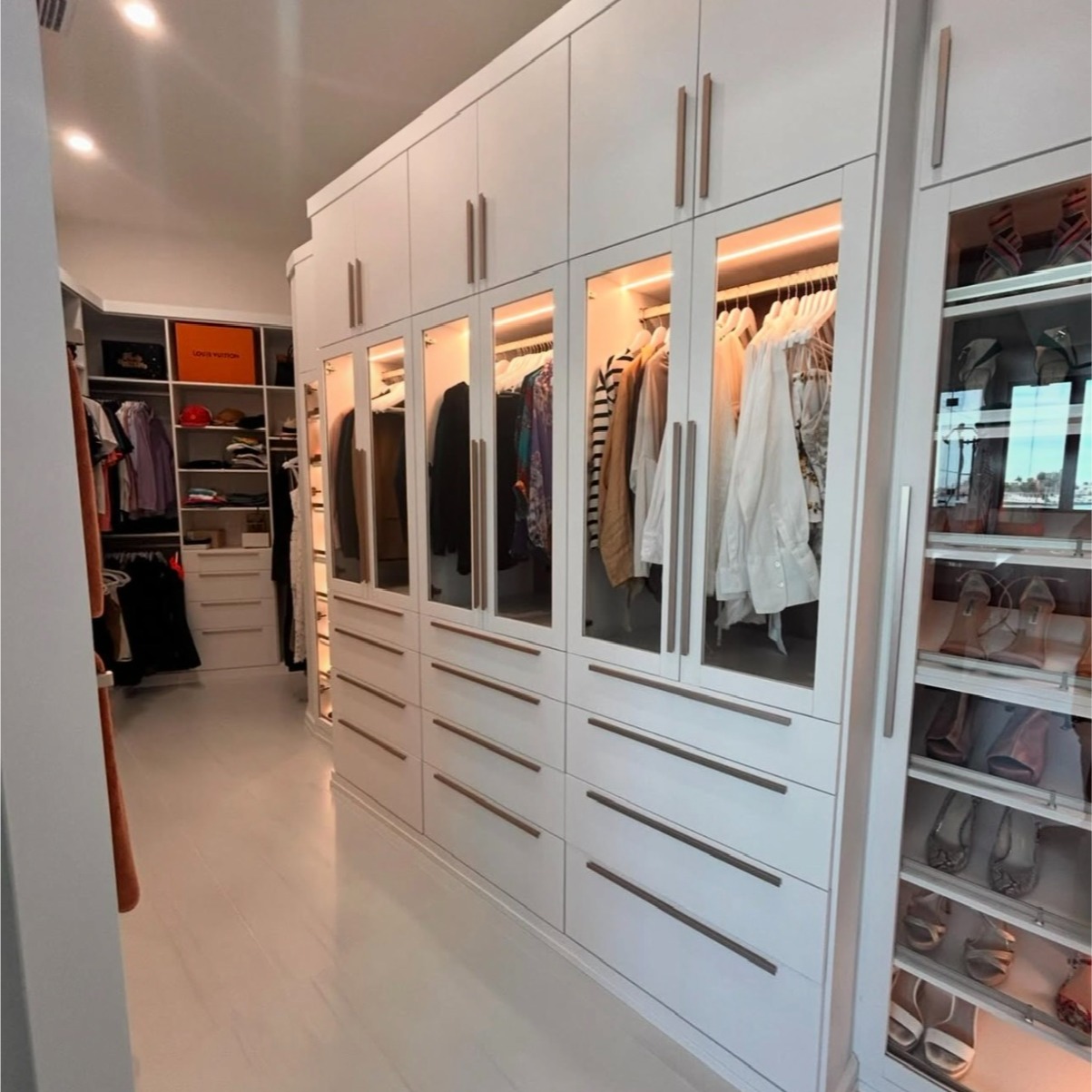 Elegant Walk-In Closet with Glass Displays and Paxton Hardware Accents Walk-in closet with a matte finish, Siena backing, glass display cabinets, strip LED lighting, and Paxton hardware.