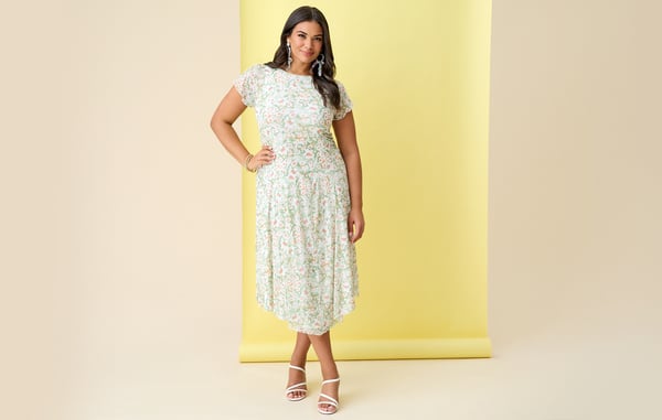Woman is wearing a plus size light green floral midi dress and is posing with one hand on her hip in front of a yellow background