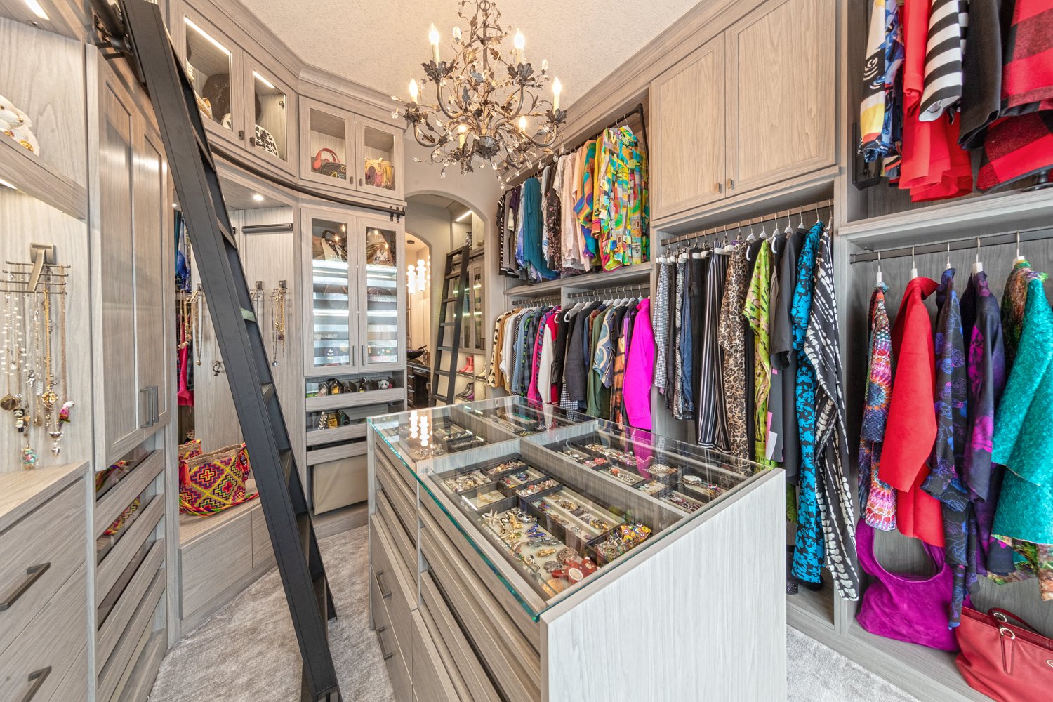 Spacious Walk-In Closet Full of Details Custom walk-in closet designed in a light gray finish. This closet features a center island, double and single hang, glass doors, lighting, a bench, ladder for easy access, and drawers for clothing and accessories.