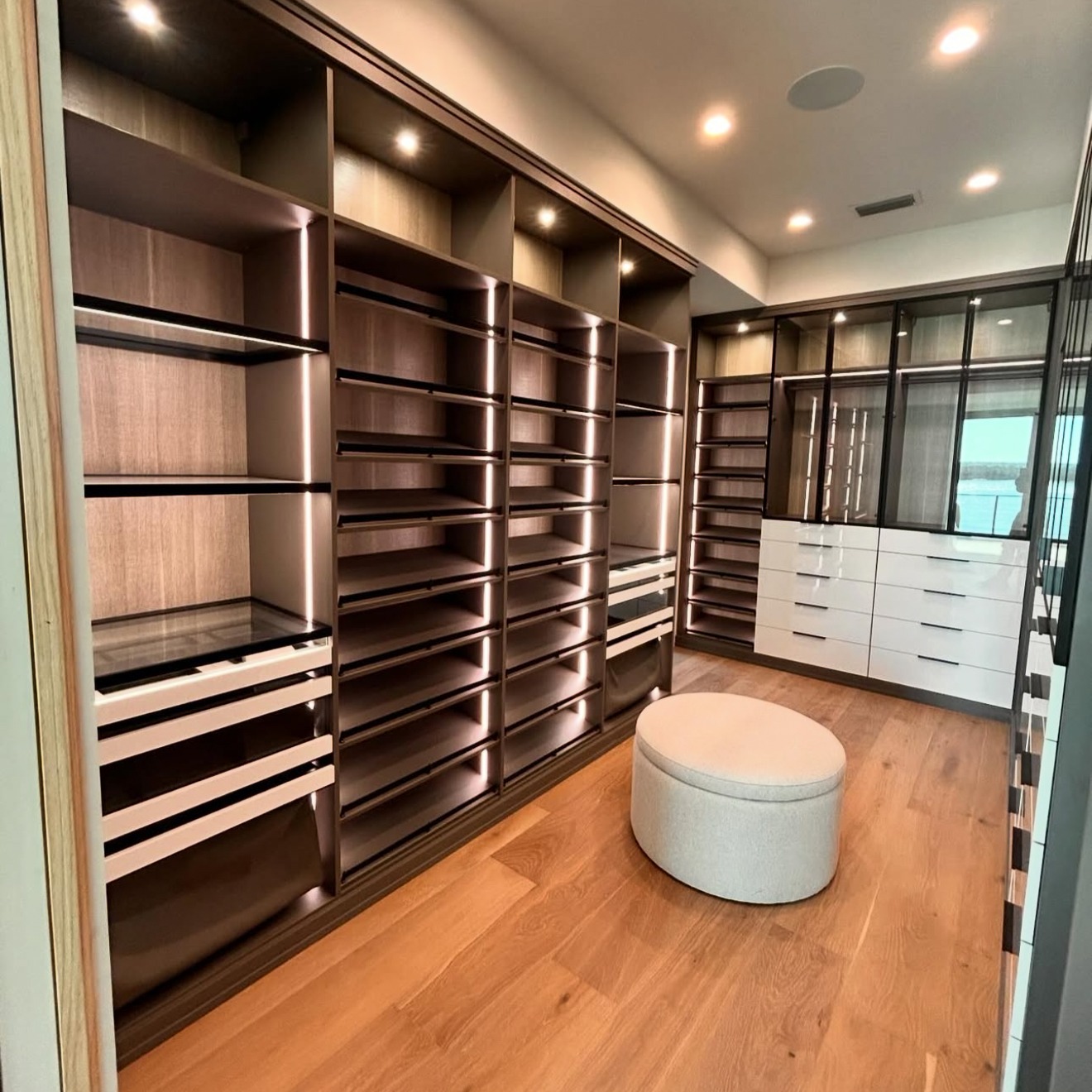 Sleek Walk-In Closet with Modern Finishes and Custom Lighting Walk-in closet featuring Shipshaw finish, Pietra backing, Element Designs aluminum frame doors with finger pulls and black hardware, high gloss drawer fronts, Everstyle drawers, and illuminated by strip and puck LED lighting.