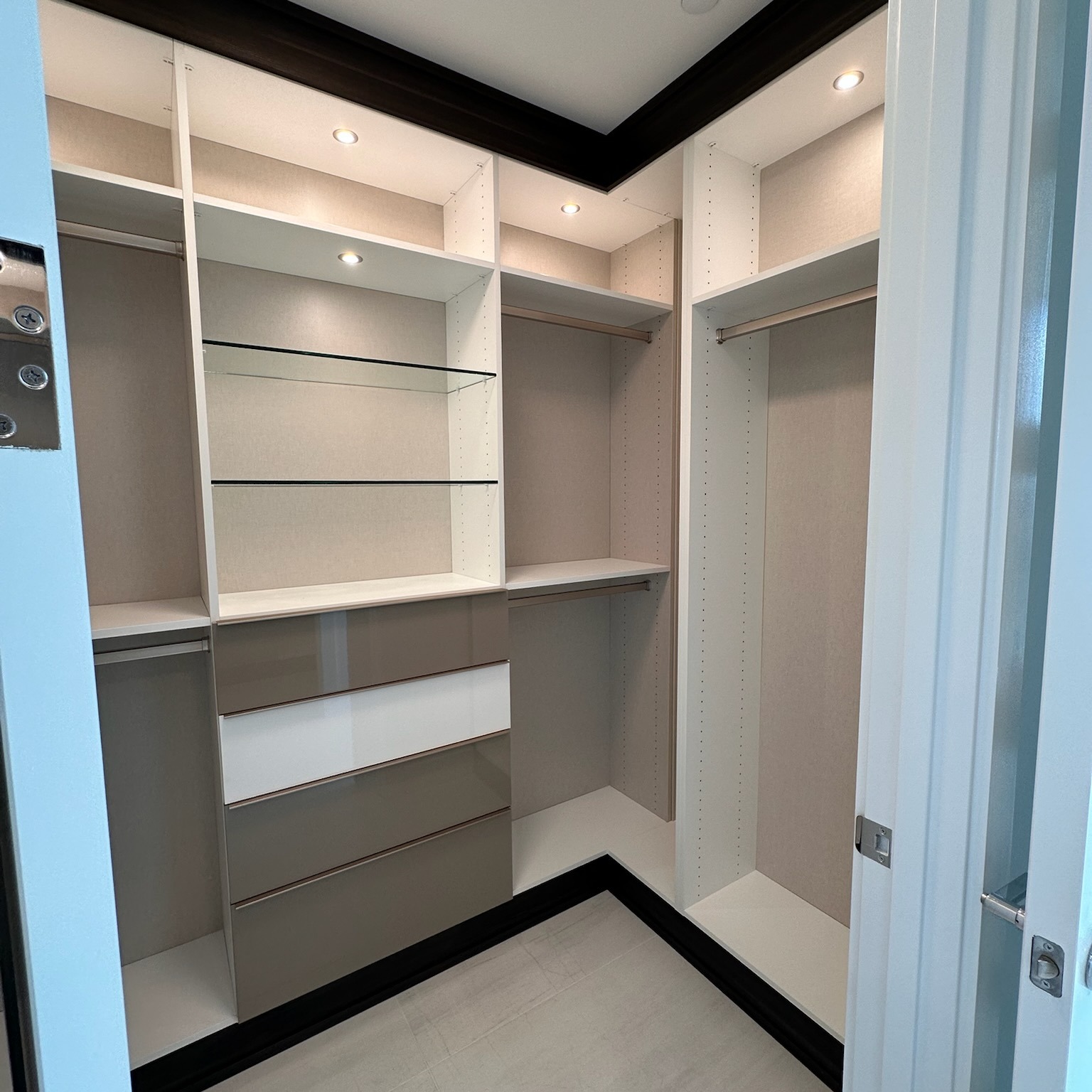 A Master Closet That Marries Style & Function Custom master closet with Dove partitions and shelving, Linen backing, Cappuccino trim, matte nickel hardware, tempered glass shelves, high-gloss Soho slab drawer fronts in Stone Grey and Bright White, recessed matte aluminum puck lights, and Engage laundry hamper in Slate with matte nickel accents.