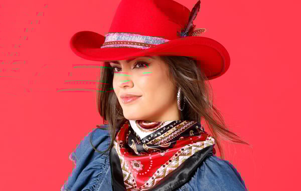 Woman is wearing a red cowboy hat and a red and black paisley scarf, and is posing in front of a red background.
