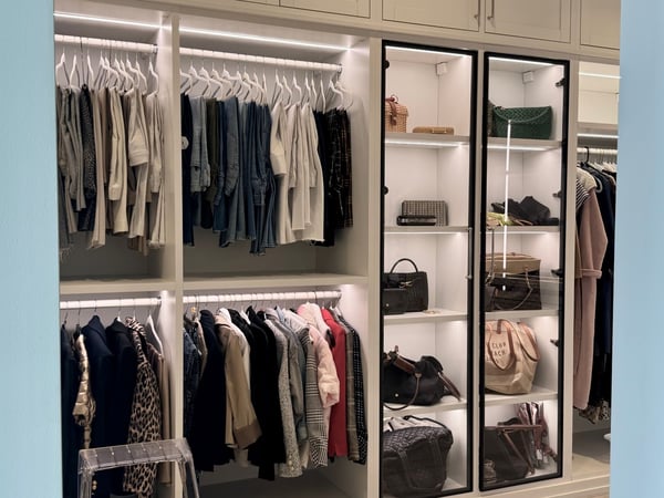 A refined walk-in closet that combines open wardrobe storage with beautifully illuminated display cabinetry for a polished, boutique-inspired feel. Luxury walk-in closet with illuminated hanging storage, glass-front display cabinets, shelving, and custom wardrobe organization.