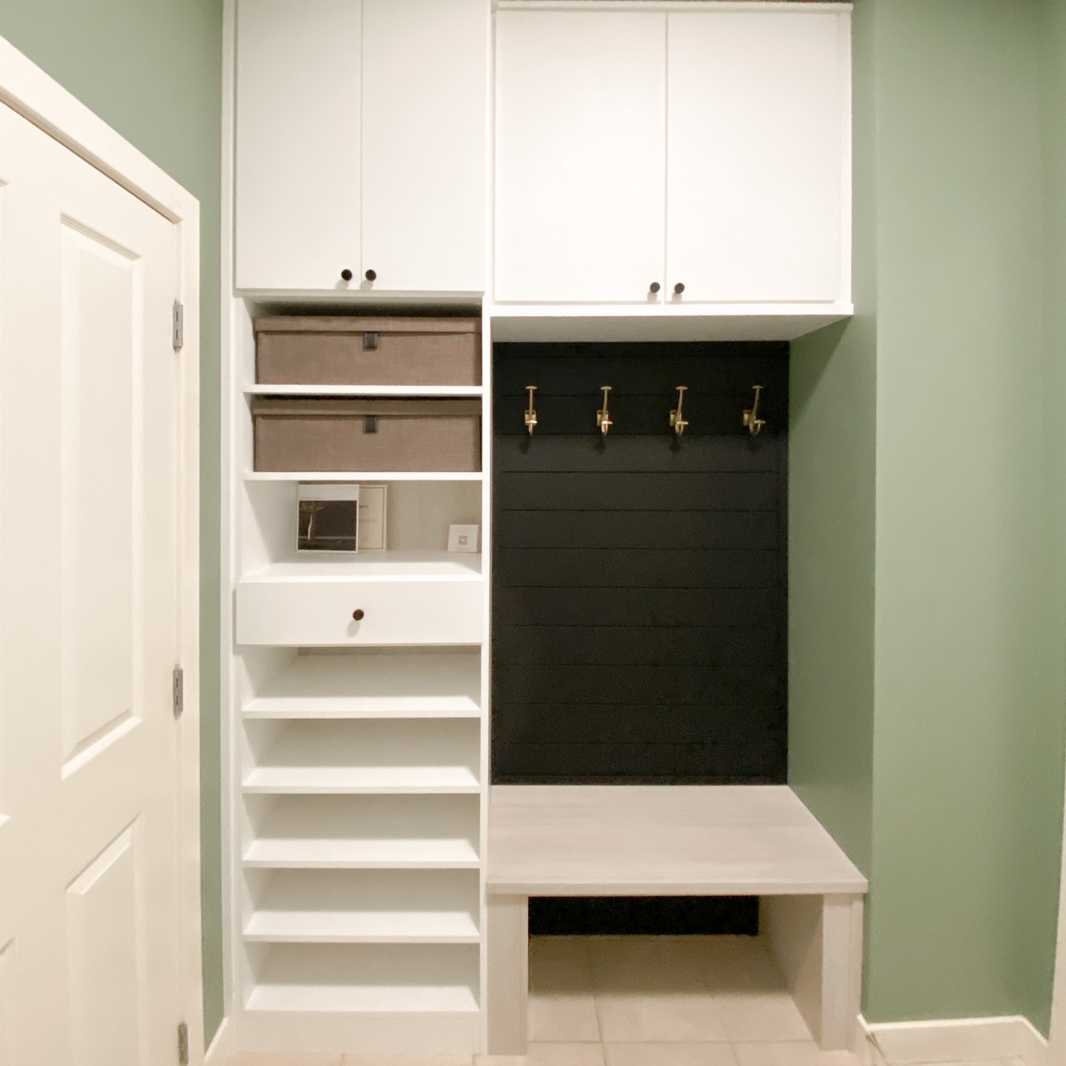 Simple But Functional Mudroom Small mudroom nook filled with upper cabinets, shoe shelves, a bench, and black shiplap with gold hooks.