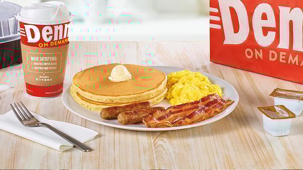 To-go cup of Denny's Coffee, a white plate containing a stack of pancakes with a dollop of butter, two sausage links, two slices of bacon and scrambled eggs, two to-go containers of syrup.