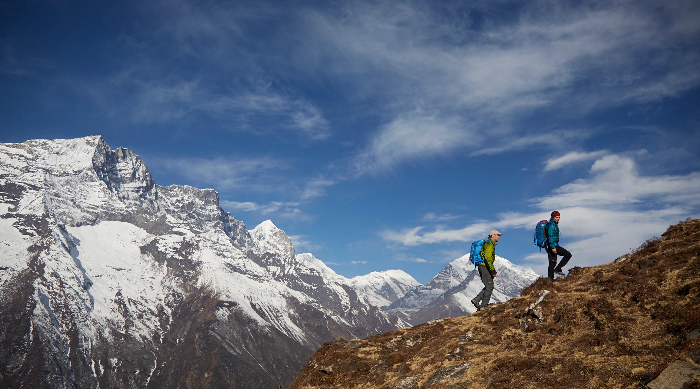 Two hikers wearing puffy jackets, hats, and large day-hike packs ascend a mountain ridge with snowy peaks behind them.