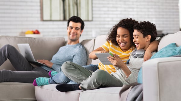 A father sits on a couch using his laptop as his children watch a movie on their tablet using COX Communications to stay connected.