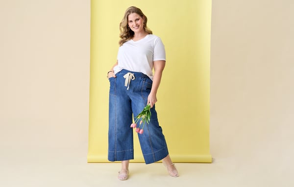 Woman is wearing a plus size white v neck top tucked into curvy wide leg cropped jeans, and is posing with one hand in her pocket and holding tulips in front of a yellow background.