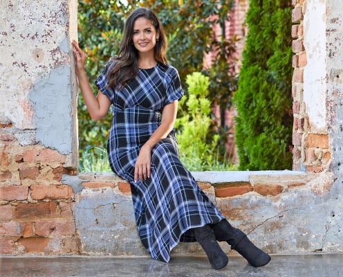 Woman wearing a blue plaid maxi dress is sitting on a brick ledge.