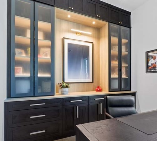 This custom office built-in by California Closets strikes the perfect balance between elegance and utility. Crafted in a rich, dark wood finish with a contrasting white oak backdrop, the design features sleek, glass-front upper cabinets with integrated lighting to showcase books, art, and personal mementos. Ample drawer and cabinet storage below offers a clutter-free workspace, while the central niche—with accent lighting and space for artwork or a monitor—adds a refined focal point. Polished hardware and seamless detailing elevate the overall aesthetic, making this workspace as beautiful as it is functional.