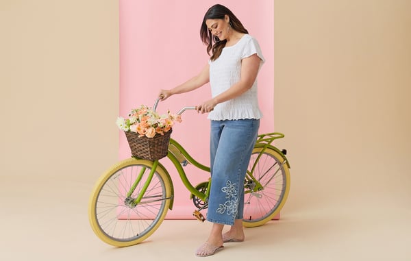 Plus size model is wearing a white blouse and wide leg cropped embroidered jeans, and is posing with a green bicycle in front of a pink and tan background.